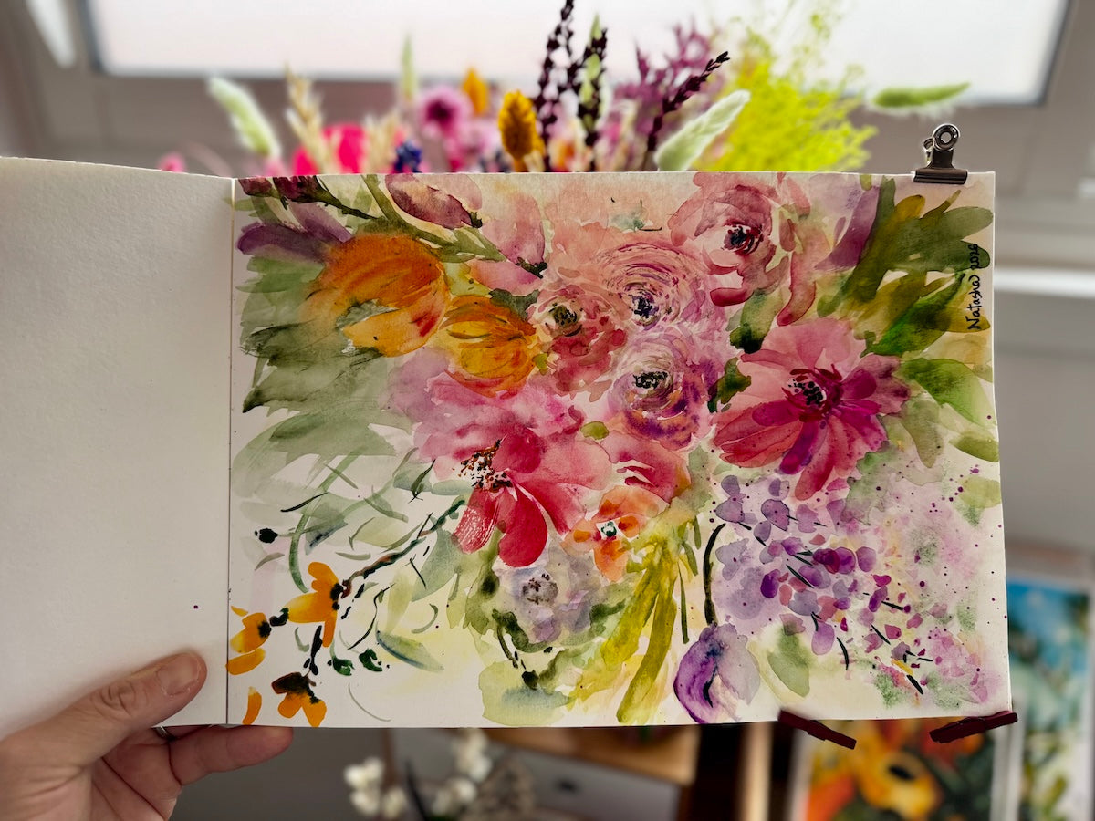 Open sketchbook held by hand, showing a loose watercolor painting of a vibrant floral bouquet with pink, purple, yellow, and orange flowers, soft green foliage, and expressive, fluid brushstrokes.