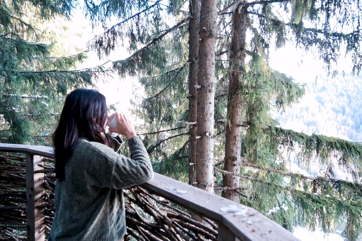 Person (Natasha Gonzalez) standing on a wooden forest balcony, drinking from a cup while overlooking snow-dusted evergreen trees and a mountain landscape. Boutique de Moments by Natasha Gonzalez. MOMENTOS number 21
